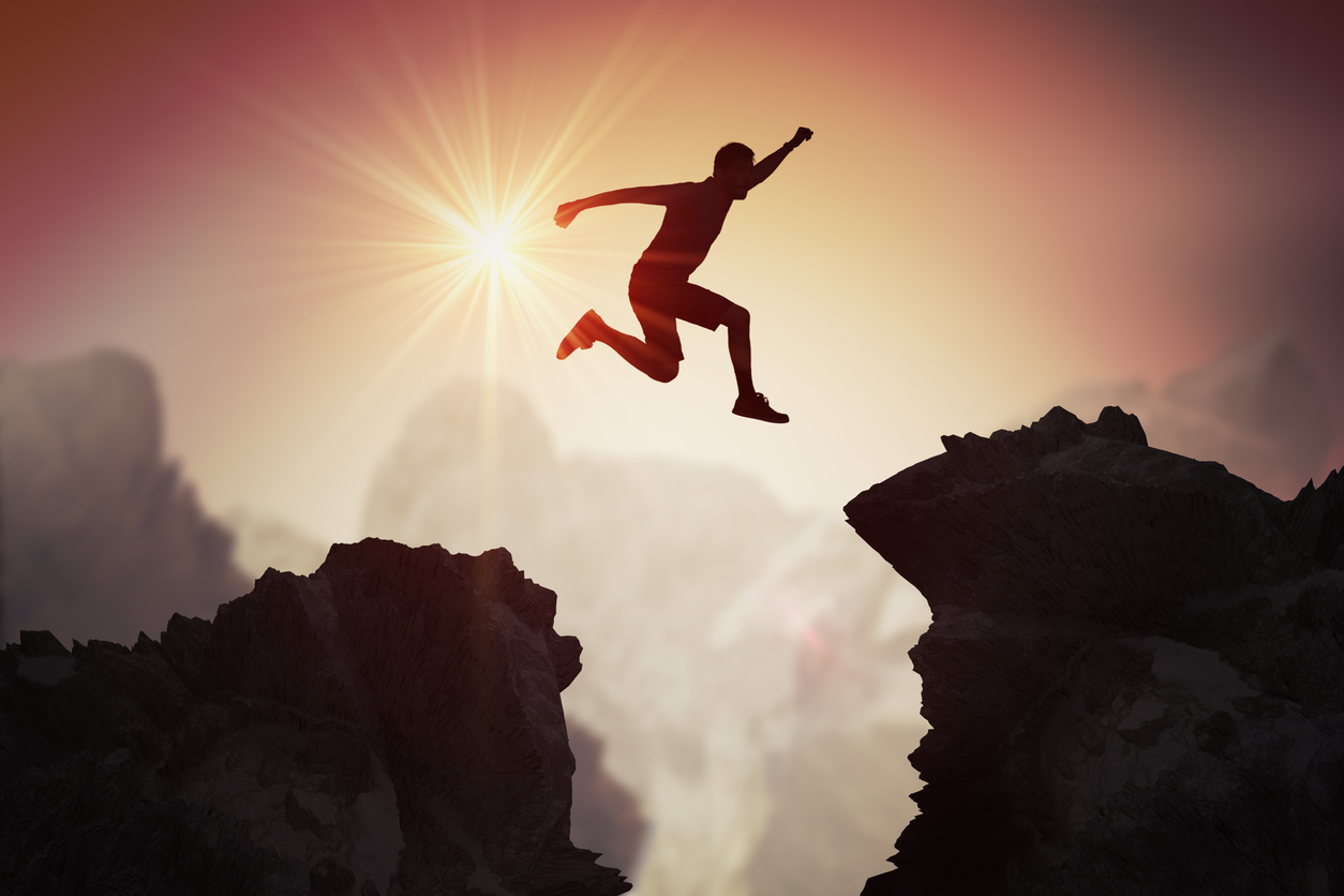 Home silhouette of young man jumping over mountains and cliffs at sunset.